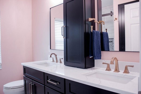 Bathroom vanity with two sinks, bronze faucets, and a tall black medicine cabinet between dark wood cabinets; pink walls and blue towels visible.